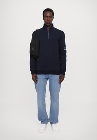 Sudadera de forro polar azul marino con cuello de media cremallera, bolsillo frontal tipo canguro y detalle de logo en la manga, combinada con jeans azul claro y zapatillas blancas.