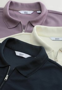 Trois polos texturés pliés avec fermetures éclair en lavande, crème et noir, chacun étiqueté « NEXT Regular Fit » sur le col.