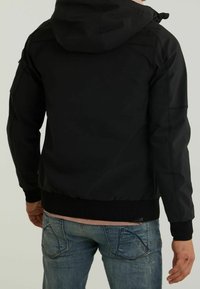 Homme portant une veste noire à capuche et un jean bleu, vu de dos sur un fond clair uni.