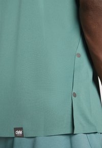 Teal athletic top made from textured fabric with side slits and gray dot accents. Features a black label stating "ciele Athletics."