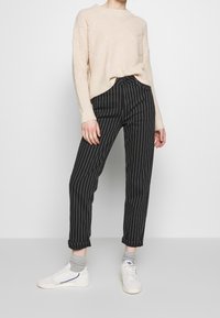 Beige stickad tröja med ett diamantmönster, kombinerad med svarta randiga byxor och vita sneakers. Synliga uppvikt ärmar och ljusgrå strumpor.