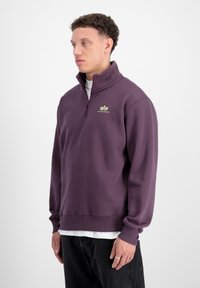 Felpa a girocollo di colore viola scuro, con collo a zip quarti, polsini e orlo a costine, e un piccolo logo verde sul lato sinistro del petto.