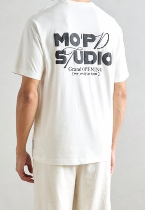 Hvid bomulds t-shirt med en afslappet pasform. Sort trykt tekst på ryggen siger "MO'PD STUDIO Grand OPENING [ses kl. 20:00]."