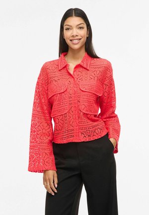 Kobieta o długich ciemnych włosach, uśmiechnięta, ubrana w jasną koralową koronkową koszulę na guziki z dużymi przednimi kieszeniami oraz czarne spodnie, stoi z ręką w kieszeni.