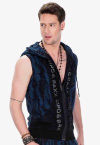 Marineblaues ärmelloses Hoodie mit einem strukturierten Muster, ausgestattet mit einem frontalen Reißverschluss und auffälligen Logoakzenten in Silber. Mit einem schwarzen Taillenbund gesäumt.