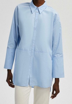 Personne portant une chemise bleu clair ample à boutons avec des poignets larges et un pantalon crème, sur un fond uni.