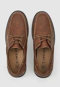 Zapatos de cuero marrón con costuras y cordones blancos. Se puede ver la puntera redondeada y una plantilla texturizada. Diseño minimalista, estilo casual.