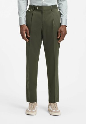 Mann, der olivgrüne plissierte Hosen und beige Sneaker trägt, vor einem schlichten weißen Hintergrund stehend.