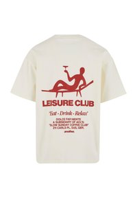 Kreminė medvilnės marškinėliai su dideliu raudonu grafiku, kuriame pavaizduota gulinčio figūra su gėrimu, tekstas „LEISURE CLUB“ ir papildomi detalės.