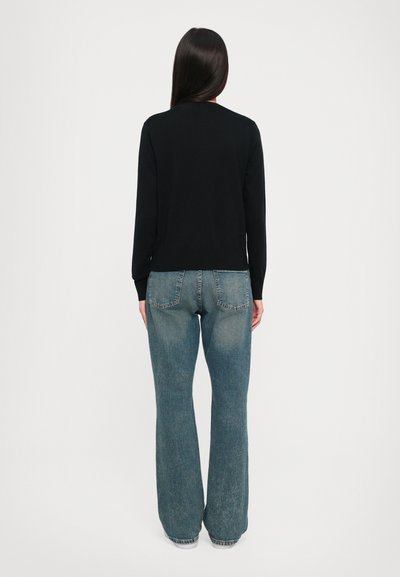 Donna con capelli lunghi e scuri, vestita con una maglietta nera a maniche lunghe e jeans blu larghi, in piedi di spalle su uno sfondo bianco.