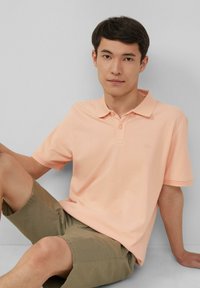 Licht perzikkleurige poloshirt met een kraag, korte mouwen en twee knopen; gecombineed met olijfgroene shorts. Gladde textuur, casual ontwerp.