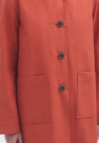 Cappotto di cotone rosso con taglio dritto, dotato di chiusura a bottoni frontale, due tasche applicate e una tasca sul petto. Bottoni scuri aggiungono contrasto.
