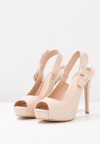Sandali con tacco a piattaforma in vernice nuda con punta aperta, dotati di cinturino alla caviglia e decorazione a fiocco, caratterizzati da un sottile tacco alto e una superficie liscia.