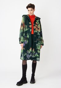 Green patterned wool coat with floral designs, featuring a hood, large buttons, and contrasting mustard accents. Worn over an orange turtleneck.