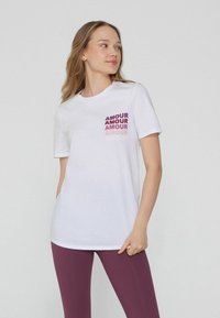 Camiseta de algodón blanca con un gráfico de la palabra "AMOUR" en diferentes tonos de rosa y morado en el lado izquierdo; combinada con leggings morados.