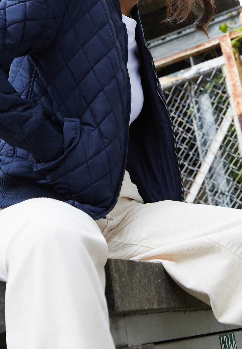 Navy quilted bomber jacket with ribbed cuffs and side pockets, paired with cream corduroy pants. Background features a chain-link fence.