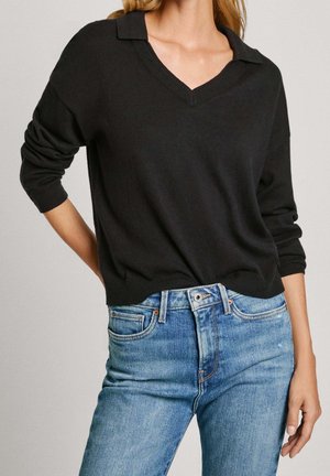 Vrouw die een zwarte lange mouw V-hals trui en blauwe high-waist jeans draagt, met haar hand op haar heup tegen een effen achtergrond.