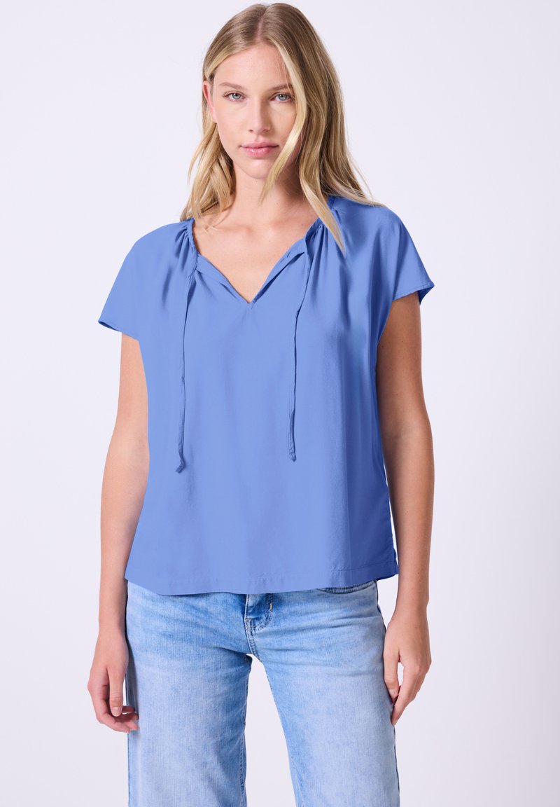Maglietta azzurro chiaro a maniche corte con scollatura a V, dettagli con cordoncino e vestibilità comoda, realizzata in un tessuto liscio. Abbinata a jeans in denim chiaro.
