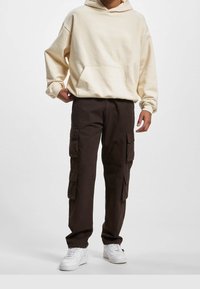 Crème hoodie met een voorzak en een capuchon met koord, gecombineerd met donkerbruine cargobroek met meerdere zijzakken. Witte sneakers maken de look compleet.