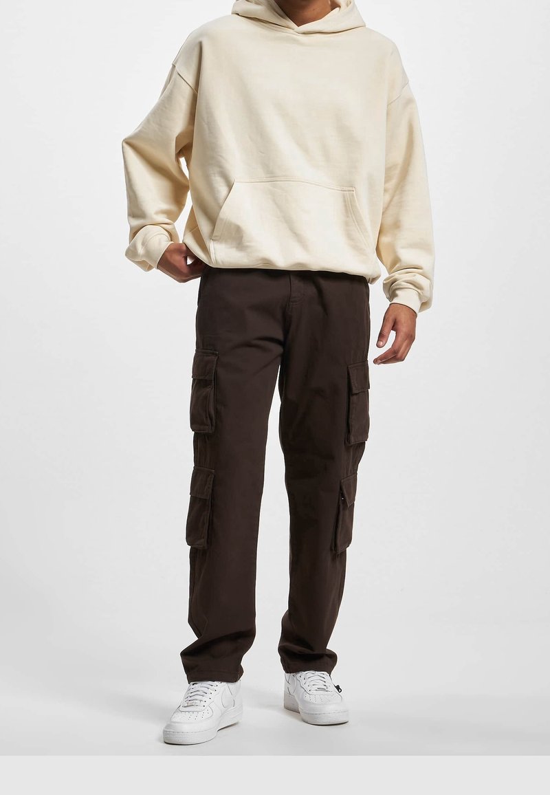 Crème hoodie met een voorzak en een capuchon met koord, gecombineerd met donkerbruine cargobroek met meerdere zijzakken. Witte sneakers maken de look compleet.