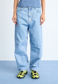 Osoba koja nosi široke svijetloplave traperice, tamnoplavu majicu i neonske žute i crne tenisice stoji na plavom tepihu.