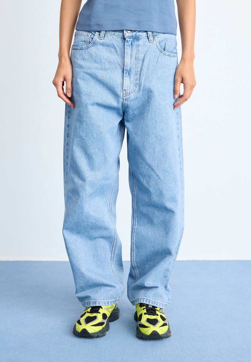 Osoba koja nosi široke svijetloplave traperice, tamnoplavu majicu i neonske žute i crne tenisice stoji na plavom tepihu.