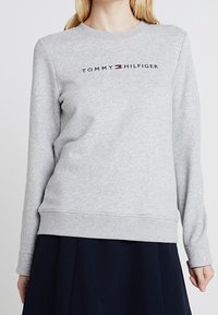 Femme portant un sweat à capuche gris clair de Tommy Hilfiger avec le texte du logo centré sur la poitrine et une jupe sombre.