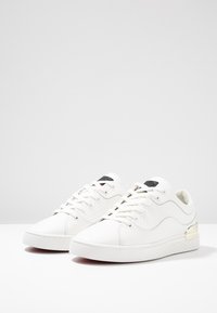 White sneakers with a textured side panel, flat laces, and a gold-accented heel. Features a smooth leather finish and rubber sole.