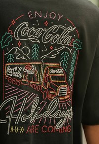 Black T-shirt featuring colorful embroidered design of a truck, trees, mountains, and holiday text with "Coca-Cola" branding.