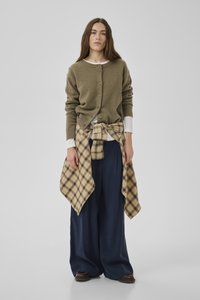 Frau trägt olivgrüne, geknöpfte Strickjacke über weißem Oberteil, karierte Hemd um die Taille gebunden, weite marineblaue Hose und braune Schuhe, steht vor einfachem Hintergrund.