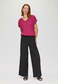 Fuchsia T-shirt met korte mouwen en een V-hals, gecombineerd met losse zwarte wijde broekspijpen. Model draagt zwarte sandalen met bandjes. Effen achtergrond.