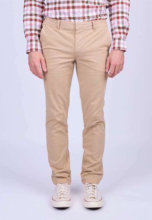 Chinos beiges avec une coupe ajustée, en tissu de coton lisse, cinq poches et passants de ceinture, associés à des baskets blanches avec des accents rouges.