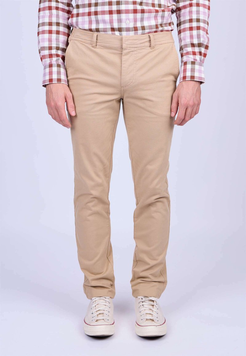 Chinos beiges avec une coupe ajustée, en tissu de coton lisse, cinq poches et passants de ceinture, associés à des baskets blanches avec des accents rouges.