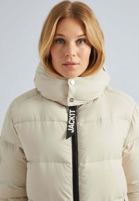 Veste beige rembourrée avec un col montant et une fermeture éclair noire, arborant un grand étiquette noire avec "JACKIT" à l'avant. Texture lisse.