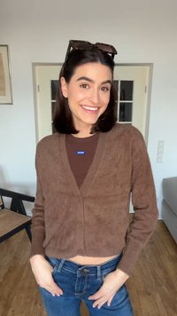 Smilende kvinne med mørkt hår iført brun cardigan, solbriller på hodet og blå jeans, står i et lyst rom med tregulv og møbler.