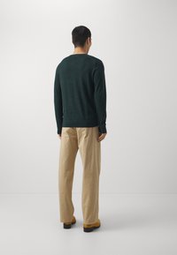 Man vänd bort mot kameran, iklädd en mörkgrön långärmad tröja, beiga vida byxor och tan mockaskor, mot en helt vit bakgrund.