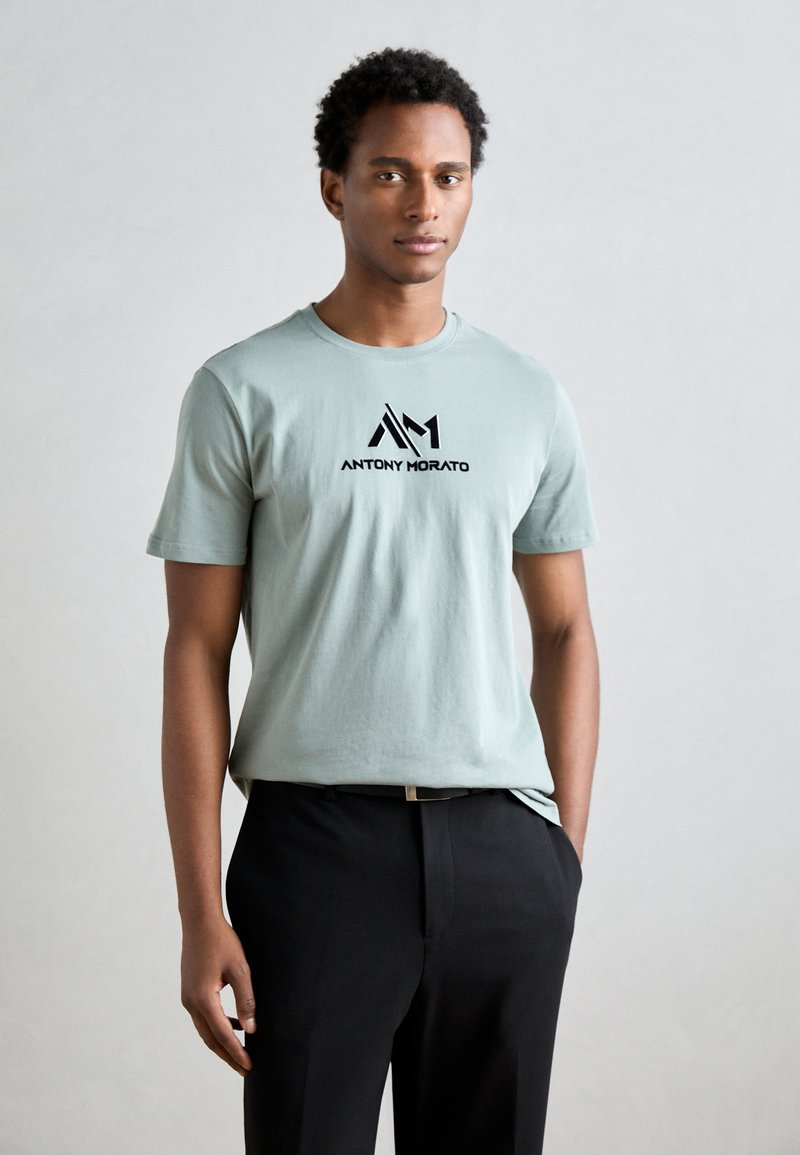 Lichtgroene katoenen t-shirt met een zwart logo "AM" en "ANTONY MORATO" op de voorkant, gecombineerd met zwarte broek.