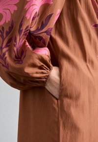 Braunes Kleid mit pinkem Blumenmuster an den Ärmeln. Besitzt geraffte Ärmelbündchen und eine Seitentasche. Glatter, strukturierter Stoff.