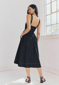 Robe en denim avec un corsage ajusté, des bretelles larges et une découpe dans le dos. Jupe évasée avec des accents de couture visibles. Bottines brunes jusqu'aux chevilles avec des sangles.