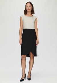 Robe bicolore avec un dessus crème à manches courtes et une jupe portefeuille noire. Le tissu est lisse avec une silhouette ajustée et une longueur jusqu'aux genoux.