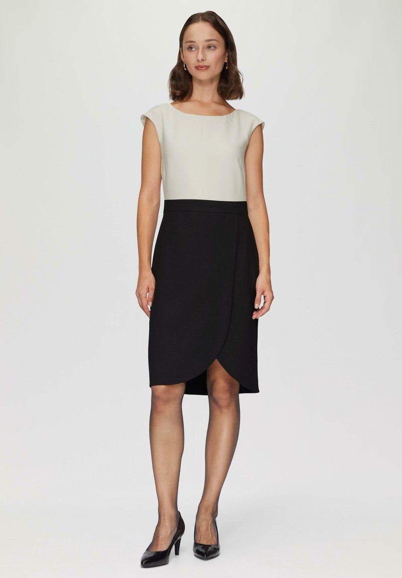 Robe bicolore avec un dessus crème à manches courtes et une jupe portefeuille noire. Le tissu est lisse avec une silhouette ajustée et une longueur jusqu'aux genoux.