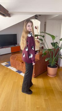 Bordowy sweter z białymi i czarnymi wzorami yin-yang, połączony z czarnymi spodniami i czarnymi butami; wnętrze z rośliną i meblami.