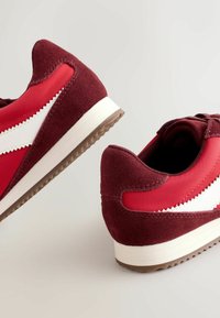 Červené sportovní boty vyrobené z semiše a syntetických materiálů, s bílým cik-cak proužkem, kulatou špičkou a texturovanou gumovou podrážkou.