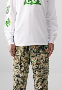 Langermet hvit skjorte med grønne grafiske trykk, kombinert med camo cargo-bukser med brune, grønne og svarte bladmønstre og lommer.
