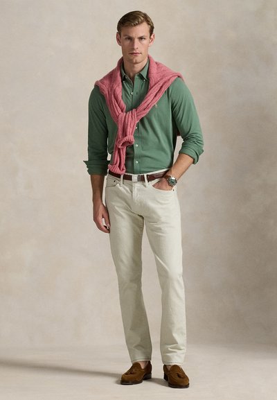 Mann trägt grünes Hemd mit Knöpfen, hellbeige Hose, braune Loafers, rosa Pullover über die Schultern gelegt, steht vor einfarbigem Hintergrund.