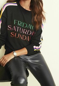 Zwarte trui met kleurrijke tekst "VRIJDAG ZATERDAG ZONDAG" en gestreepte mouwen. Model draagt glanzende zwarte leren broek en een zilveren horloge.
