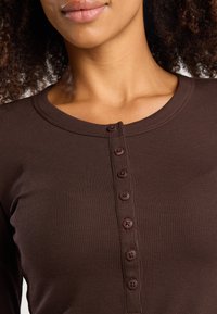 Bruine geribbelde langeklauw top met een ronde halslijn, voorzien van zes bijpassende bruine knopen aan de voorkant. Soepele textuur en getailleerd ontwerp.