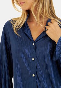 Camisa de satén azul marino con textura ondulada, cuello puntiagudo y botones en tono dorado. Mangas largas con puños abotonados.