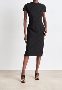 Femme portant une robe midi noire ajustée à manches courtes et des talons ouverts avec des brides noires, brunes et crème, debout sur un sol clair.