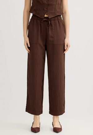 Mujer vistiendo pantalones de tiro alto, sueltos, color marrón oscuro con bolsillos frontales y cordón, combinados con tacones marrones a juego con punta puntiaguda.